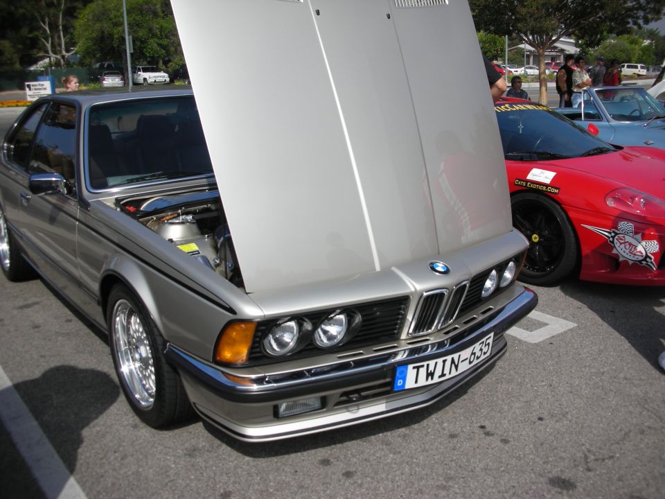 Silver BMW E24 coupe, hood open, parked next to a red Ferrari. License plate reads TWIN-635