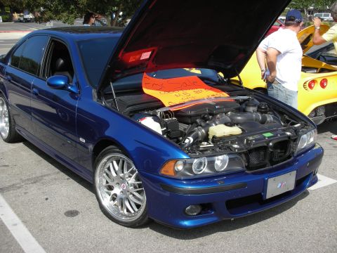 1998 Ford Crown Victoria - A blue BMW M5 sedan is parked with its hood open, revealing the engine and a 'For Sale' sign.