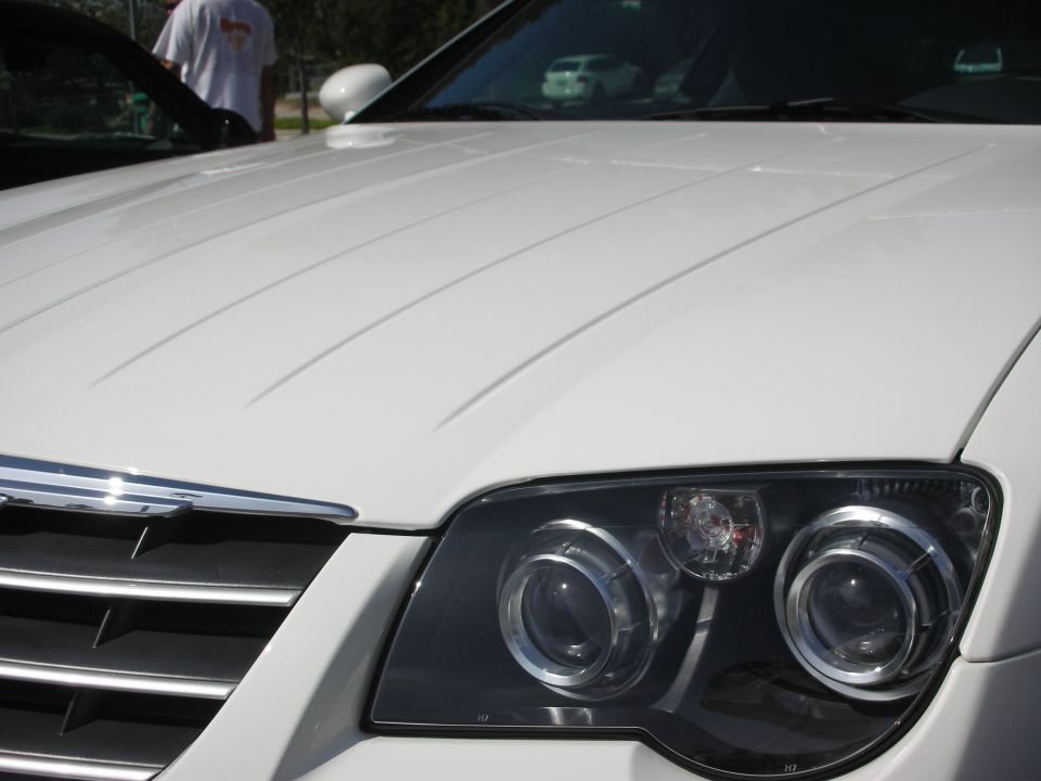 Front end of a white Chrysler Crossfire, showing the headlights and grille. Another car and a person are visible in the background