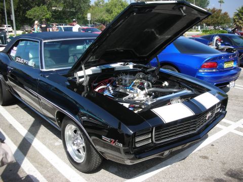 1998 Ford Crown Victoria - A black 1969 Chevrolet Camaro Z/28 is parked with its hood open, revealing the engine. Another car is visible in the background.
