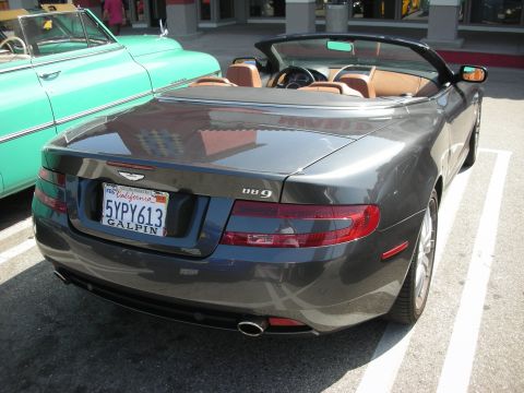 1998 Ford Crown Victoria - A dark gray Aston Martin DB9 convertible is parked in a lot next to a vintage teal car.