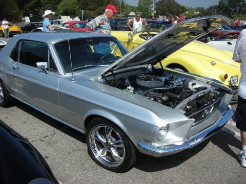 1998 Ford Crown Victoria - A silver 1967 Ford Mustang coupe is displayed with its hood open at a car meet, its engine and chrome wheels.