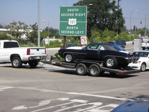 1998 Ford Crown Victoria - A black 1969 Chevrolet Camaro is transported on a trailer, with a white pickup truck in front and a highway sign in the background.