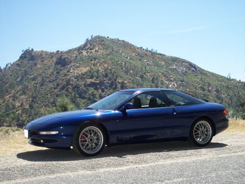 1995 Ford Probe - Metallic blue Ford Probe GT hatchback parked on a gravel shoulder in front of a scrub-covered rocky peak, shot low from the front three-qua…