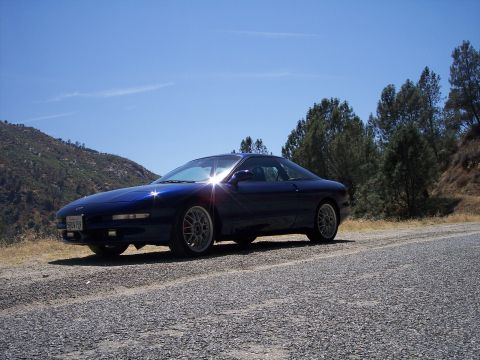1995 Ford Probe - A blue 1995 Ford Probe is parked on the side of a road with trees and a hill in the background.