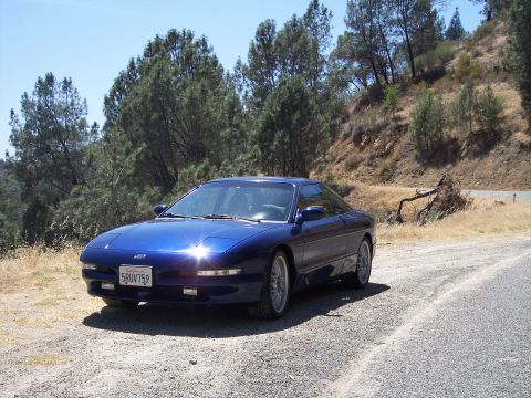 1995 Ford Probe - Dark blue Ford Probe hatchback coupe parked on a gravel turnout beside a pine-covered hillside, shot from the front three-quarter driver's…
