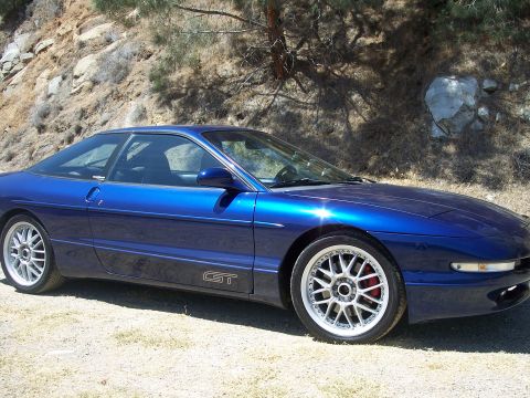 1995 Ford Probe - Metallic blue Ford Probe GT hatchback shot from a low front three-quarter angle on a dirt shoulder beside a rocky, brush-covered hillside.…