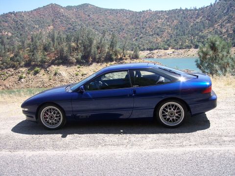 1995 Ford Probe - Full side profile of a metallic blue Ford Probe GT hatchback coupe parked on a gravel shoulder above a green lake with brush-covered hills…