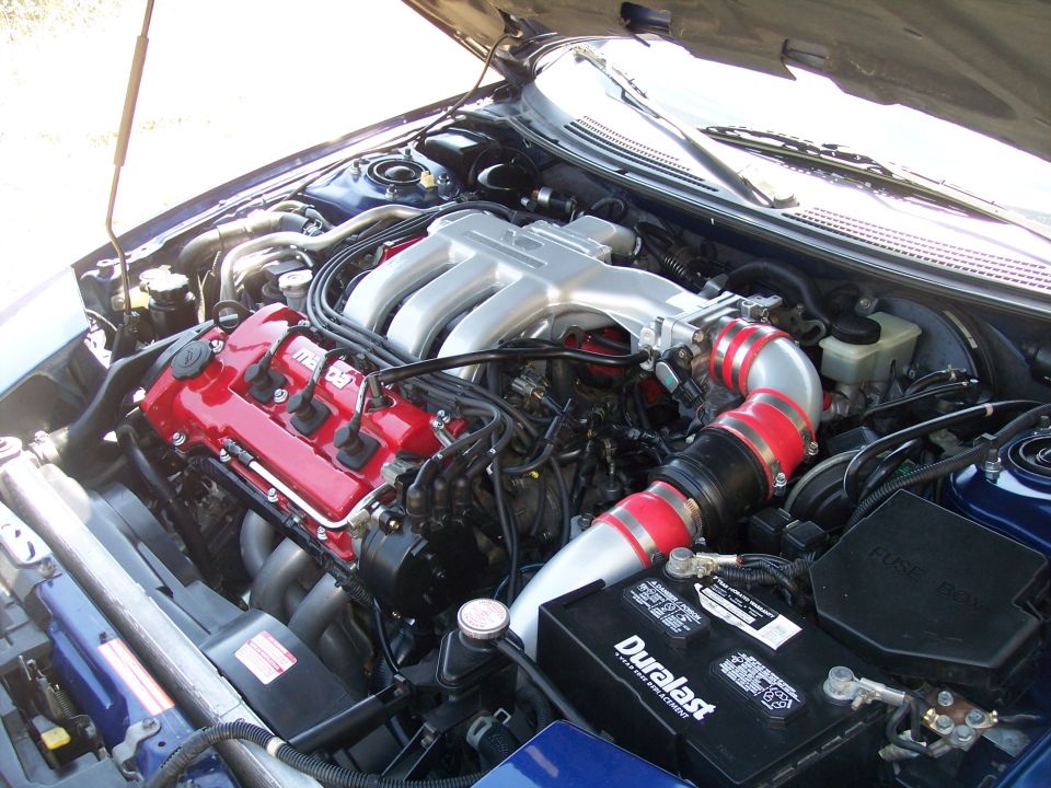 Mazda V6 engine bay with red valve cover, silver plenum, intake pipe, battery and fuse box