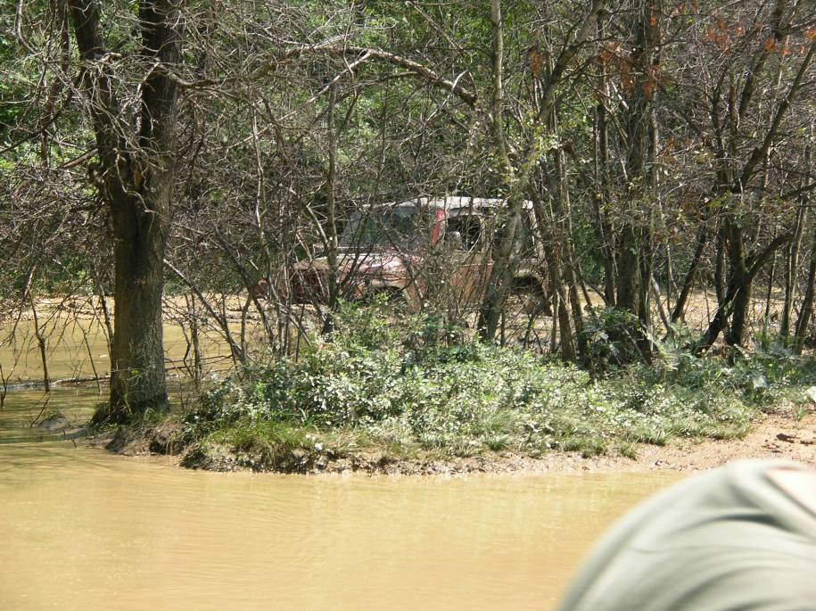 Red SUV partially obscured by trees, driving through muddy water