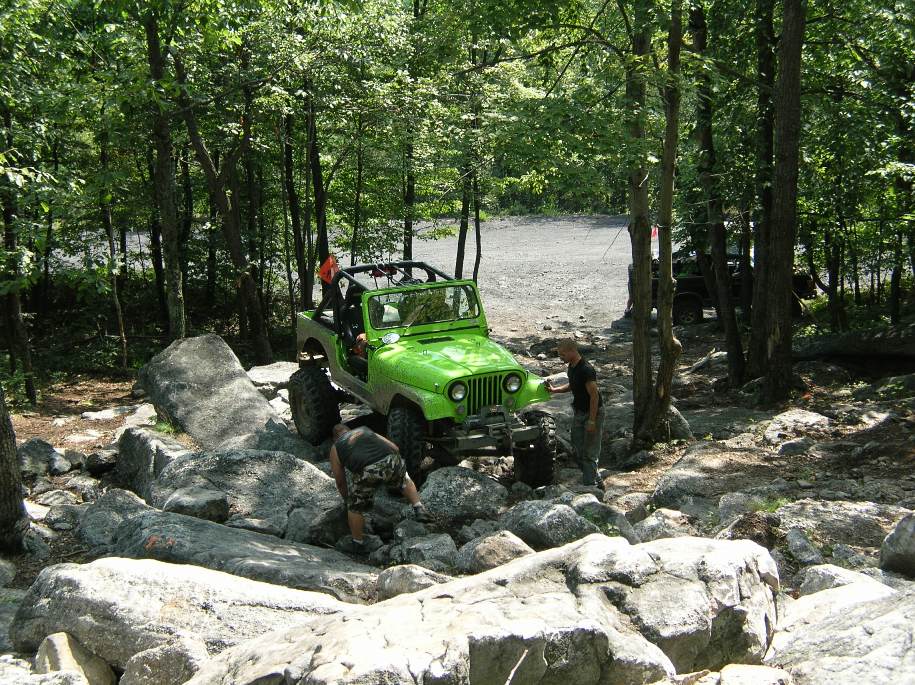 Green Jeep Wrangler navigating large rocks on a wooded trail; another vehicle partially visible in background