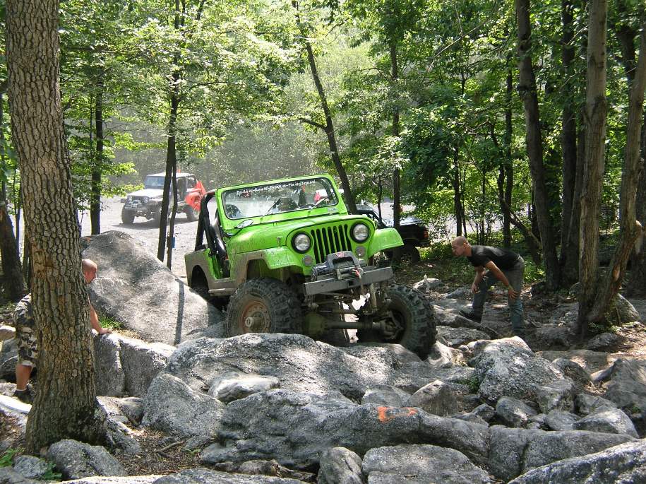 Green Jeep Wrangler navigating a rocky trail, with another Jeep and an orange vehicle in the background