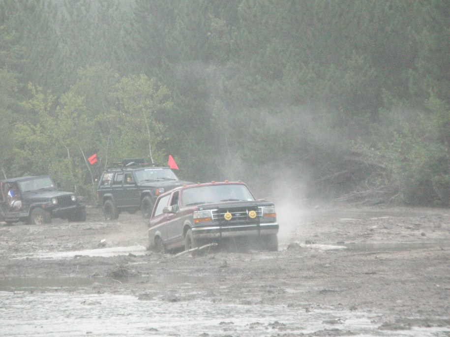 Three off-road vehicles navigating a muddy track, with the lead vehicle being a red and silver Ford Bronco