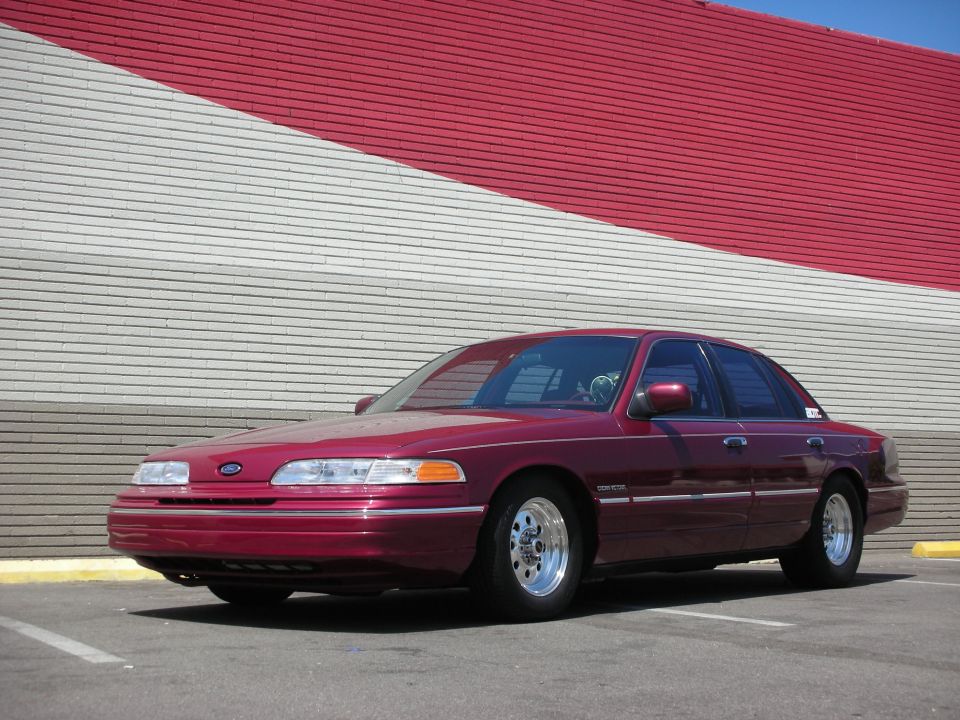 Maroon Ford Crown Victoria parked in front of a red and white striped wall. It has aftermarket wheels