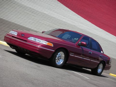 1992 Ford Crown Victoria - A maroon 1992 Ford Crown Victoria sedan is angled towards the camera in front of a building with red and white striped siding.