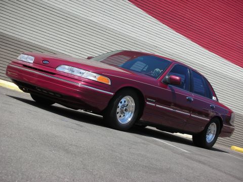 1992 Ford Crown Victoria - A maroon 1992 Ford Crown Victoria sedan is angled towards the camera in front of a building with a red and white striped facade.