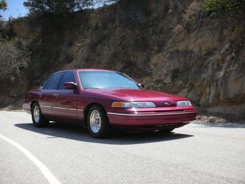 1992 Ford Crown Victoria - This maroon sedan is parked on the side of a road with a rocky incline in the background.