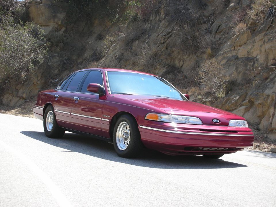 Maroon Ford Crown Victoria parked on a road with a rocky hillside behind it