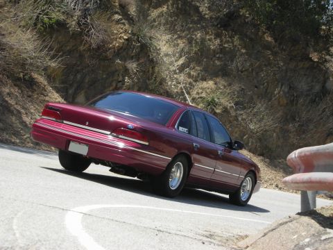 1992 Ford Crown Victoria - A maroon 1992 Ford Crown Victoria is captured from the rear as it navigates a curve on a scenic road.