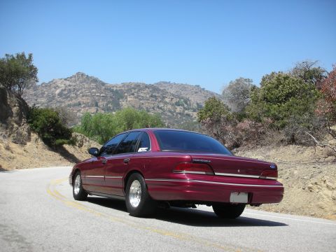 1992 Ford Crown Victoria - A maroon 1992 Ford Crown Victoria sedan is seen from the rear on a winding road with mountains in the background.