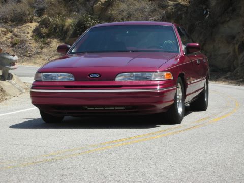 1992 Ford Crown Victoria - A maroon 1992 Ford Crown Victoria is shown from the front on a two-lane road with a rocky hillside in the background.