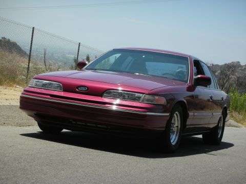 1992 Ford Crown Victoria - A maroon 1992 Ford Crown Victoria is parked on a road with a chain-link fence and dry brush in the background.