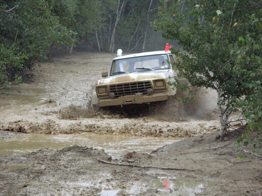 Tan off-road vehicle with a prominent grille driving through a muddy track, trees and foliage in the background