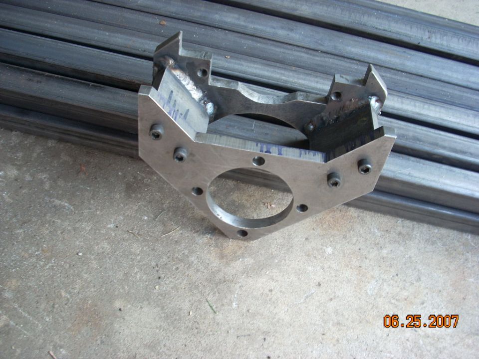Metal bracket with six bolt holes and a central circular opening, welded construction, on concrete floor with metal bars in background
