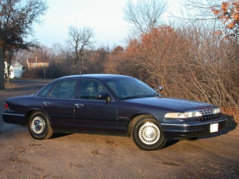 1995 Ford Crown Victoria - A 1995 Ford Crown Victoria sedan is seen from the front 3/4 angle on a dirt road with trees in the background.