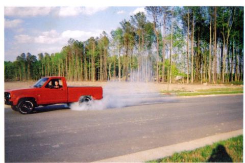 1989 Ford Mustang - A red pickup truck performs a burnout on a paved road with trees in the background.