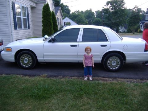 2003 Ford Crown Victoria - A white 2003 Ford Crown Victoria sedan is parked on the side of the road with a young girl standing next to it.