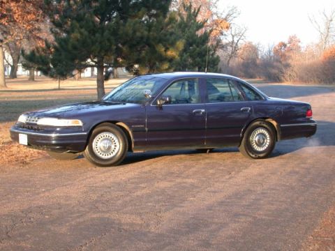 1995 Ford Crown Victoria - A dark blue 1995 Ford Crown Victoria is shown from the side, parked outdoors with trees in the background.