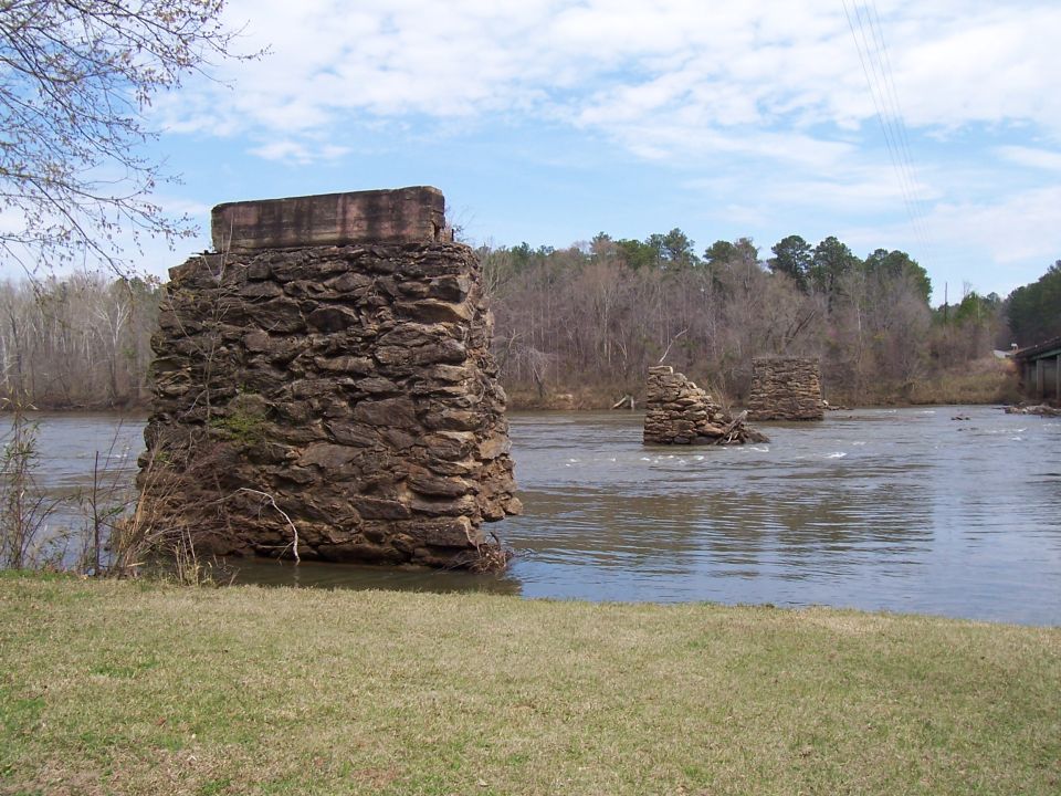 Ruined stone bridge pilings stand in a river with trees on the far bank under a cloudy sky