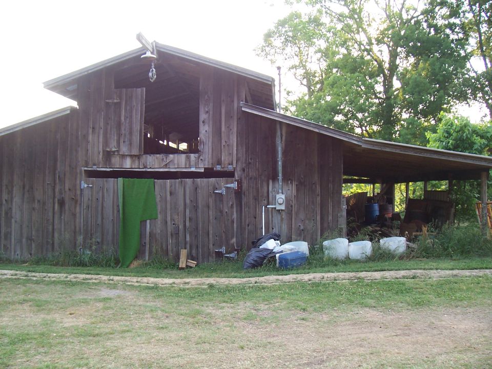 Wooden barn with open loft, green tarp hanging, and covered lean-to area with stored items