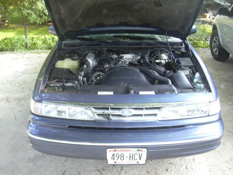 1996 Ford Crown Victoria - Front view of a blue Ford Crown Victoria with its hood open, revealing the engine compartment. A second vehicle is visible to the right.