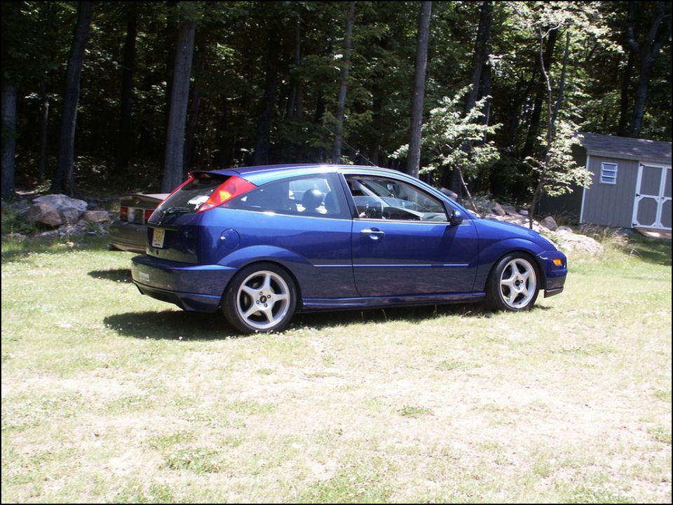 Blue hatchback parked on grass with trees and a shed in the background