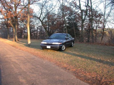 1995 Ford Crown Victoria - A dark 1995 Ford Crown Victoria is parked on the side of a road with trees in the background.