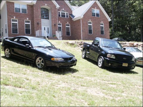 2001 Mercury Grand Marquis - Two black vehicles, a Ford Mustang coupe and a Ford F-150 pickup truck, are parked on a grassy lawn in front of a large brick house.