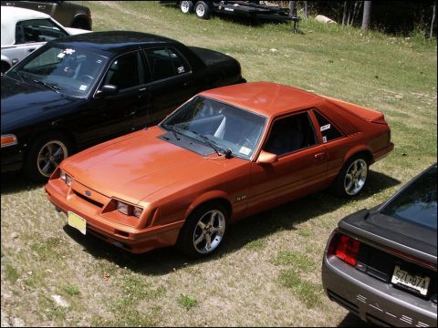 2001 Mercury Grand Marquis - An orange 1985 Ford Mustang coupe is parked next to other vehicles at an outdoor gathering.
