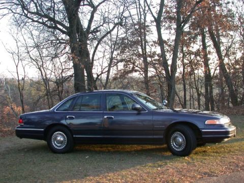 1995 Ford Crown Victoria - Side view of a dark blue 1995 Ford Crown Victoria sedan parked on grass with trees in the background.