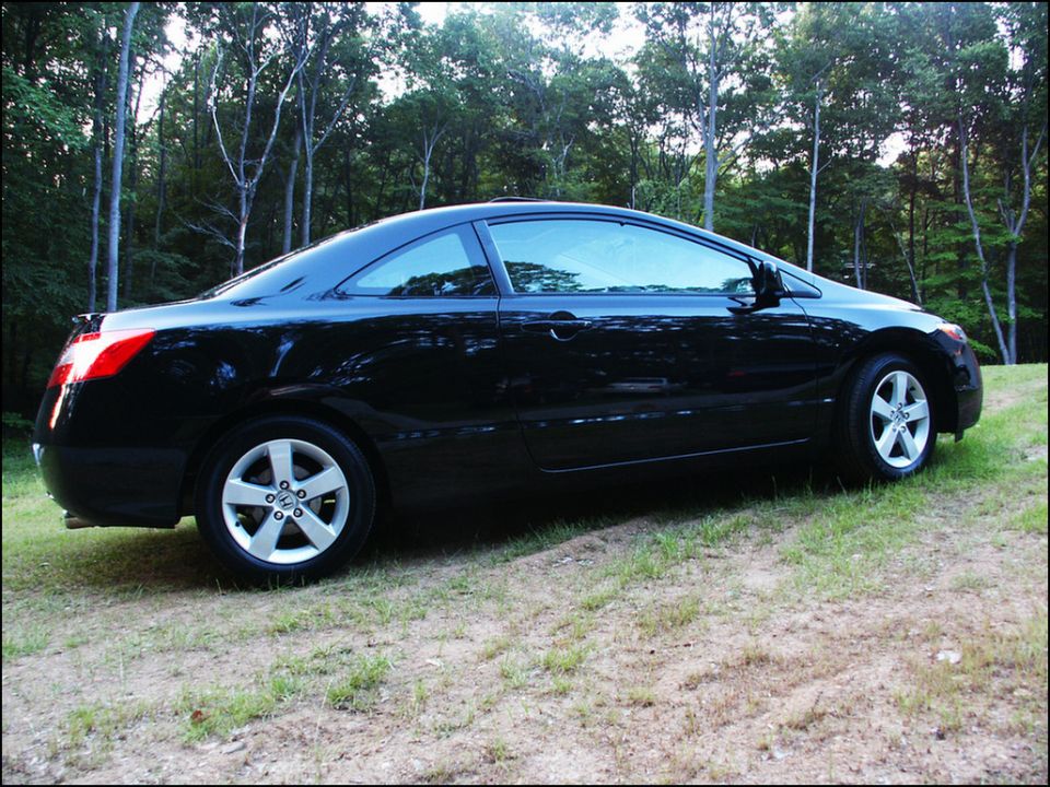 Black coupe parked on a grassy slope with trees in the background. Has a spoiler and five-spoke wheels