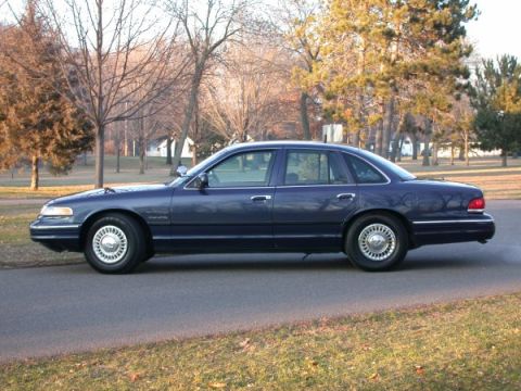 1995 Ford Crown Victoria - A dark blue 1995 Ford Crown Victoria sedan is parked on the side of a road with trees in the background.