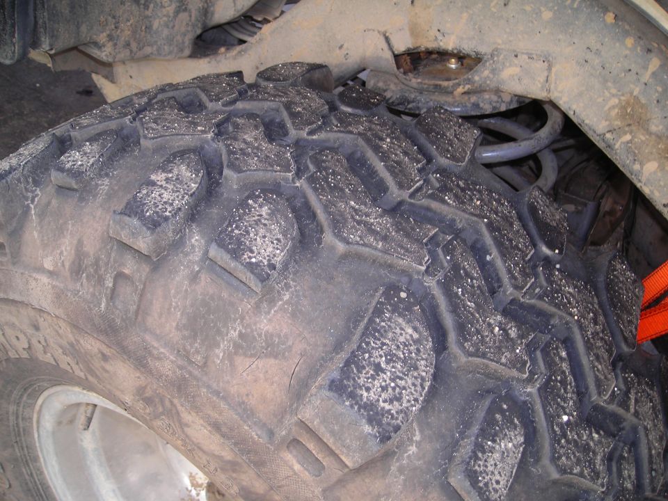 Close-up of a knobby off-road tire with visible dirt and white specks, showing suspension components and an orange strap