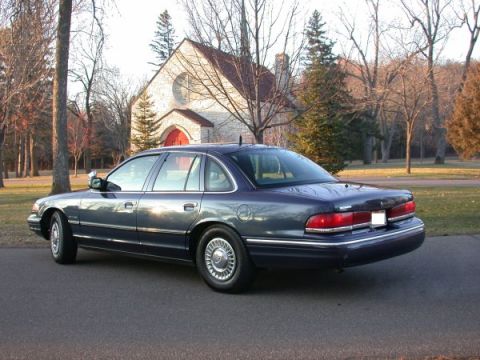 1995 Ford Crown Victoria - This 1995 Ford Crown Victoria is shown in a rear three-quarter view, parked on a paved surface with a building and trees in the background.