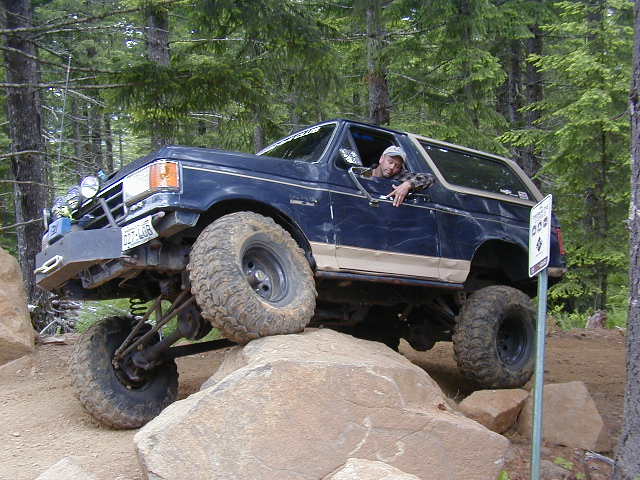 Blue and tan lifted Ford Bronco crawling boulders with driver leaning out the window