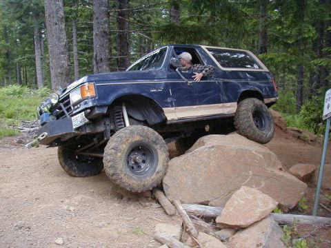 1994 Ford Explorer - This lifted 1990s era SUV, likely a Ford Bronco, tackles a rocky trail with its driver looking on.