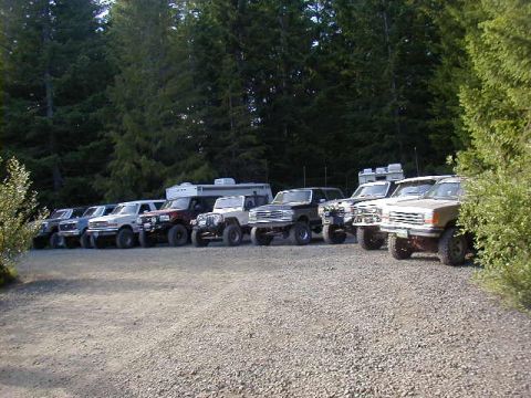 1994 Ford Explorer - A line of SUVs and trucks are parked on a gravel lot, with a dense forest in the background.
