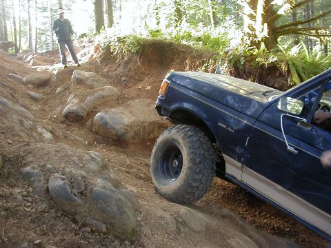 1994 Ford Explorer - A dark blue pickup truck with aggressive tires tackles a challenging, rocky off-road path in a wooded area.
