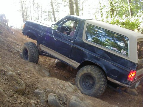 1994 Ford Explorer - A blue Ford Bronco navigates a steep, rocky incline on a wooded trail.