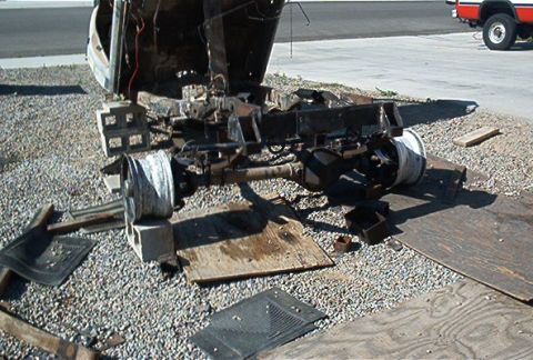 1982 Toyota 4Runner - The chassis and drivetrain of a vehicle are disassembled and resting on gravel, with a red truck visible in the background.